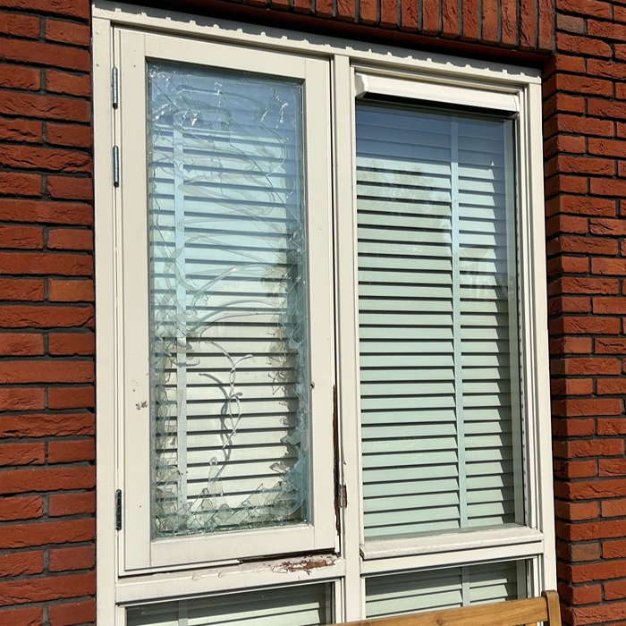 Een wit kozijn in een rode bakstenen muur, waarvan de ruit in de linkerdeur volledig is verbrijzeld met barsten in het glas.
