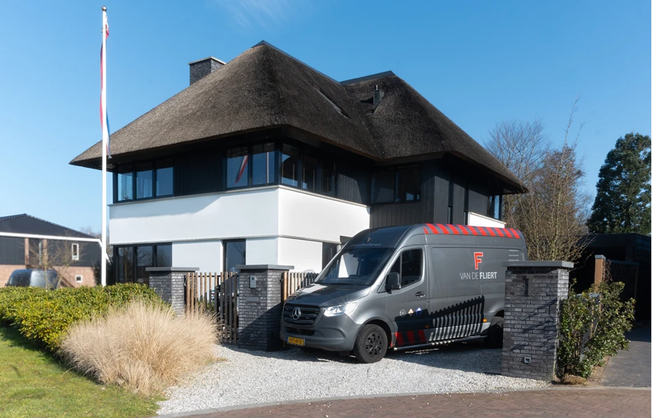 Een grijze bestelbus van het bedrijf Van de Fliert staat geparkeerd op de oprit van een moderne, luxe villa met een rieten dak.