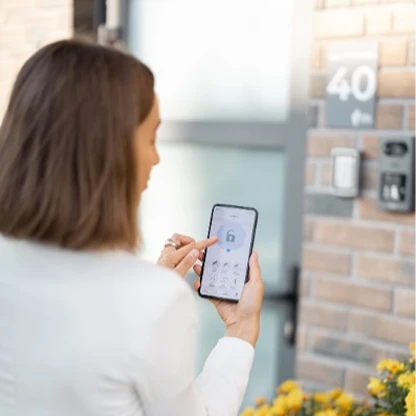 Een vrouw die via een app op haar smartphone een slim deurslot of beveiligingssysteem bedient bij de voordeur van een woning.
